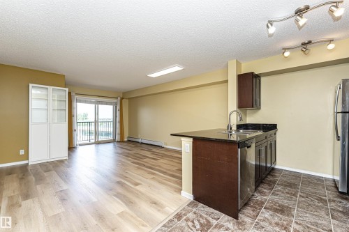 Open-concept living area featuring wood-finish flooring, a kitchen with dark cabinetry and granite-look countertops, stainless steel appliances, and a sliding glass door leading to a balcony - 404 12045 22 Avenue, Edmonton, AB 