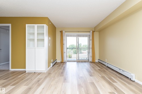 Spacious living area featuring wood-finish flooring, a built-in white cabinet with glass-front uppers, and a sliding glass door leading to a balcony - 404 12045 22 Avenue, Edmonton, AB 
