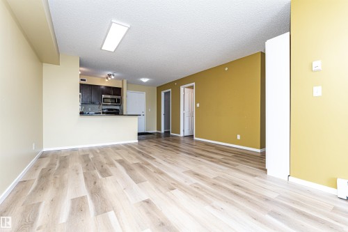 Spacious open-concept living area featuring light wood-finish flooring, a kitchen with dark cabinetry, stainless steel appliances, a breakfast bar, and recessed lighting - 404 12045 22 Avenue, Edmonton, AB 