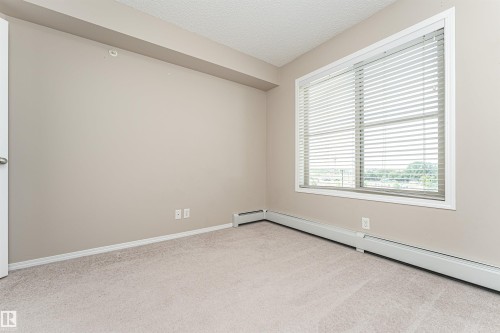 Carpeted interior room featuring neutral wall paint, white trim, baseboard heating, and a large window with horizontal blinds - 404 12045 22 Avenue, Edmonton, AB 