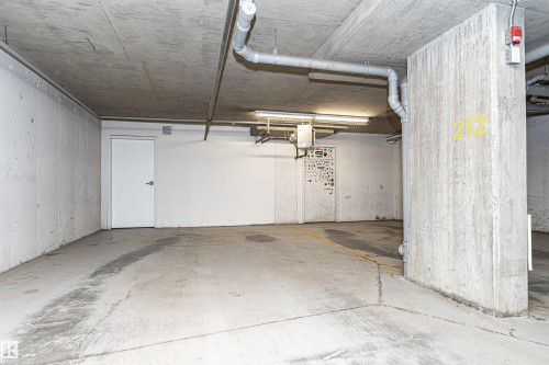 Concrete parking space featuring exposed ceiling pipes, fluorescent lighting, and a white access door - 404 12045 22 Avenue, Edmonton, AB 