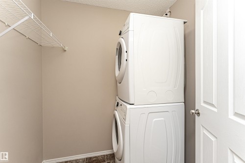 Dedicated laundry area featuring a stacked washer and dryer, wire shelving, textured ceiling, light neutral wall paint, and patterned floor tiling - 404 12045 22 Avenue, Edmonton, AB 