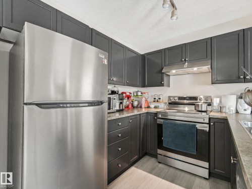 Kitchen featuring dark cabinetry, stainless steel appliances, corner sink, and wood-finish flooring - 18 230 Edwards Drive, Edmonton, AB 