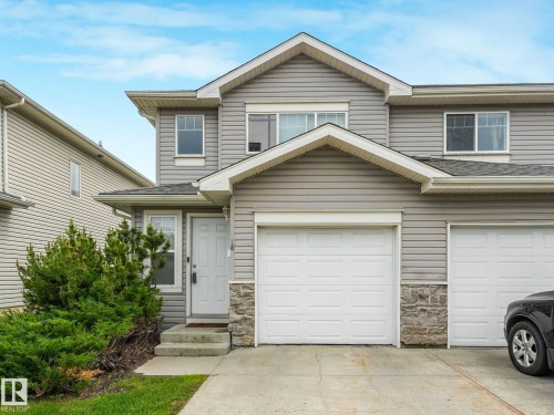 Grey siding exterior with a white front door, stone accents, attached garage, and concrete driveway - 18 230 Edwards Drive, Edmonton, AB 