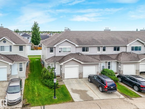 Townhome exterior featuring light gray siding, dark shingle roofing, white garage doors, and stone facade accents - 18 230 Edwards Drive, Edmonton, AB 