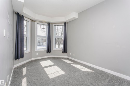 Room with a multi-pane bay window, neutral gray walls, light gray carpeting, and white baseboards - 211 10808 71 Avenue, Edmonton, AB 