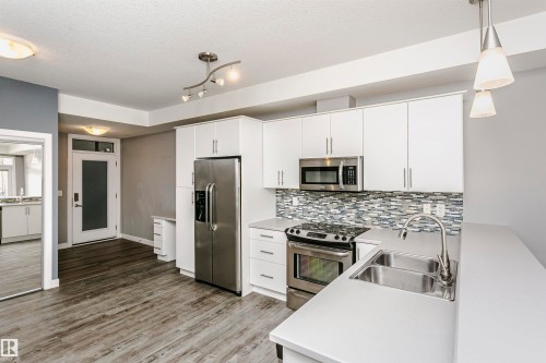 Modern kitchen featuring white cabinetry, stainless steel appliances, a mosaic tile backsplash, a double basin sink, and light-toned countertops - 211 10808 71 Avenue, Edmonton, AB 