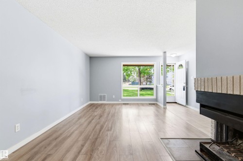 Spacious living area featuring wood-finish flooring, light gray walls, a large window, a partial view of a fireplace with a textured brick facade, and a front entry door with an arched window - 2005 Saddleback Road, Edmonton, AB - Indoor With Fireplace
