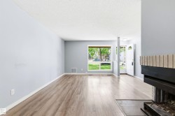 Spacious living area featuring wood-finish flooring, light gray walls, a large window, a partial view of a fireplace with a textured brick facade, and a front entry door with an arched window - 