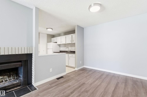 Living area featuring wood-finish flooring, a fireplace with a light brick surround and dark mantel, and a partial wall separating it from a kitchen with white cabinetry and dark countertops - 2005 Saddleback Road, Edmonton, AB - Indoor Photo Showing Other Room With Fireplace