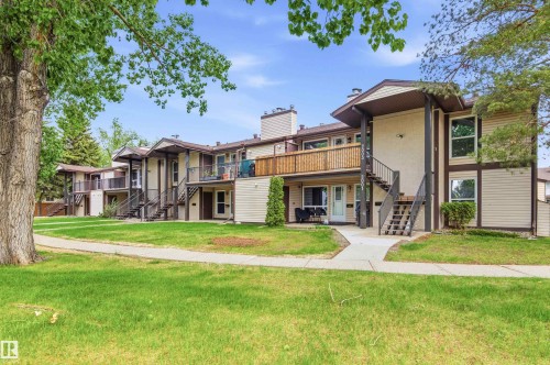 Multi-level building exterior featuring light-colored siding with dark trim, private balconies with wood railings, exterior staircases, and a green lawn with mature trees - 2005 Saddleback Road, Edmonton, AB - Outdoor