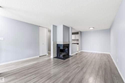 Spacious living area featuring wood-finish flooring, a brick fireplace with a dark mantel, light blue walls, and a visible kitchen area with white cabinetry - 2005 Saddleback Road, Edmonton, AB - Indoor With Fireplace