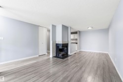 Spacious living area featuring wood-finish flooring, a brick fireplace with a dark mantel, light blue walls, and a visible kitchen area with white cabinetry - 