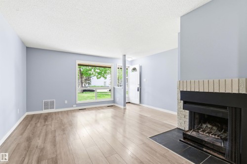 Spacious living area featuring wood-finish flooring, a fireplace with stone surround and hearth, light blue walls, and a large front-facing window - 2005 Saddleback Road, Edmonton, AB - Indoor Photo Showing Living Room With Fireplace