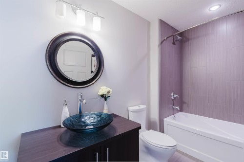 Contemporary bathroom featuring a vessel sink on a dark wood-finish vanity, a round framed mirror, a three-light vanity fixture, a toilet, and a bathtub with purple-toned vertical tiling - 4377 Mcclung Crescent, Edmonton, AB 