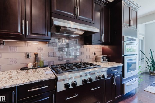 Gourmet kitchen featuring a stainless steel gas range, coordinating stainless steel ovens, rich wood cabinetry, granite countertops, and a decorative tile backsplash - 4377 Mcclung Crescent, Edmonton, AB 