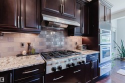 Gourmet kitchen featuring a stainless steel gas range, coordinating stainless steel ovens, rich wood cabinetry, granite countertops, and a decorative tile backsplash - 