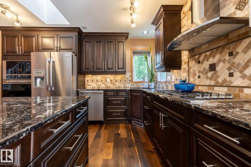 Kitchen featuring dark wood cabinetry, stainless steel appliances, speckled countertops, a gas cooktop with a stainless steel range hood, and a decorative tile backsplash - 315 Osland Place, Edmonton, AB 