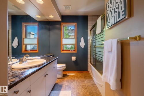Bathroom featuring a white vanity with a dark patterned countertop, dual windows with wood trim, a toilet, and a bathtub with a glass shower enclosure - 315 Osland Place, Edmonton, AB 