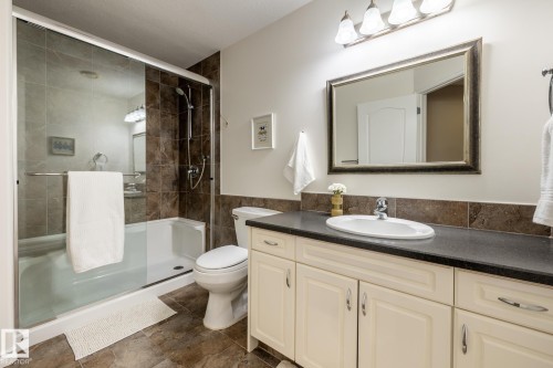 Bathroom featuring a glass-enclosed shower and tub combination with tile surround, a vanity with cream-colored cabinetry, a dark countertop, an oval undermount sink with a chrome faucet, and a rectangular framed mirror - 315 Osland Place, Edmonton, AB 