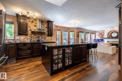 Gourmet kitchen featuring dark wood cabinetry, a granite-finish island with integrated display shelving, stainless steel appliances, a multi-tone tile backsplash, and rich wood-finish flooring - 315 Osland Place, Edmonton, AB 