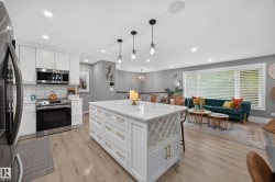 Open-concept living area featuring a large kitchen island with built-in wine storage, white cabinetry with gold hardware, stainless steel appliances, and recessed lighting - 