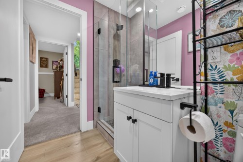 Bathroom featuring a glass-enclosed shower with tile surround, a white vanity with integrated sink and black faucet, and wood-finish flooring - 43 Burnham Place, St. Albert, AB 