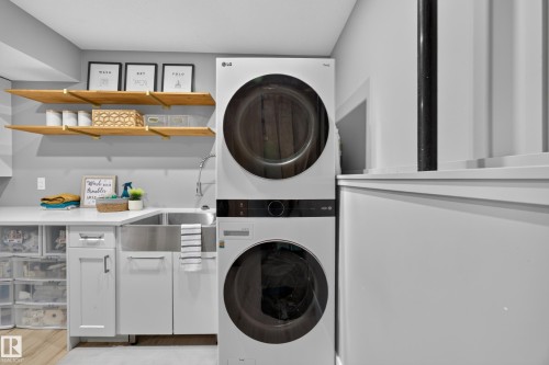 Laundry area featuring a stacked washer and dryer, a stainless steel farmhouse sink, white cabinetry with dark hardware, a white countertop, and natural wood shelving with brass brackets - 43 Burnham Place, St. Albert, AB 