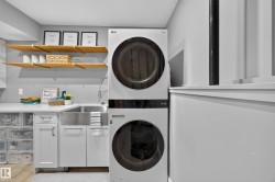 Laundry area featuring a stacked washer and dryer, a stainless steel farmhouse sink, white cabinetry with dark hardware, a white countertop, and natural wood shelving with brass brackets - 