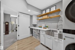 Laundry room featuring wood-finish flooring, white cabinetry, a stainless-steel farmhouse sink with a gooseneck faucet, and stacked appliances - 