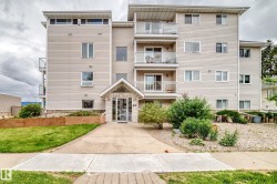 Multi-story building featuring light-toned horizontal siding, multiple balconies with glass railings, an inviting covered entrance, a concrete walkway, and landscaped garden beds - 