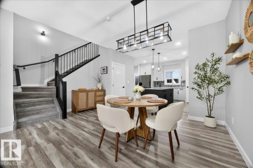 Open-concept living space featuring wood-finish flooring, a contemporary linear chandelier, and a carpeted staircase with a black metal railing - 424 Black Stone Link, Leduc, AB 