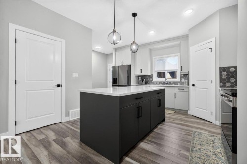 Modern kitchen featuring a dark gray island with a white countertop, light gray cabinetry, stainless steel appliances, and wood-finish flooring - 424 Black Stone Link, Leduc, AB 