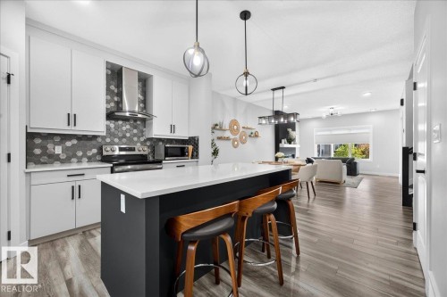 Open-concept kitchen featuring an island with a white countertop, white cabinetry with black hardware, a stainless steel range hood, a hexagonal tile backsplash, and wood-finish flooring - 424 Black Stone Link, Leduc, AB 