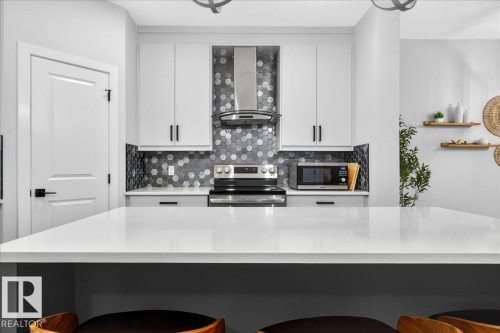 Modern kitchen featuring a white countertop island, stainless steel range and microwave, hexagonal tile backsplash, white cabinetry with black hardware, and an overhead lighting fixture - 424 Black Stone Link, Leduc, AB 