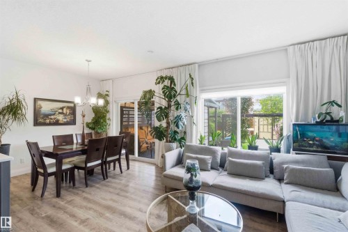 Open-concept living area with wood-finish flooring, featuring a dining space with an overhead chandelier, and a living area with large windows and a sliding glass door - 9323 223 Street, Edmonton, AB 