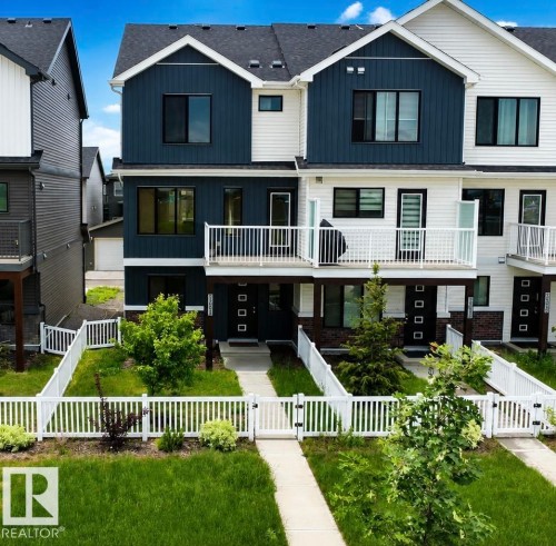 Modern multi-story townhome featuring a blue and white facade, dark-frame windows, and a white picket fence enclosing a front yard with mature landscaping - 5034 Rivers Edge Way, Edmonton, AB 