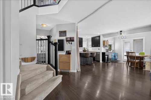 Open-concept main level featuring dark wood-finish flooring, a kitchen with dark cabinetry and white countertops, a dining area with direct outdoor access, a carpeted staircase, and a visible powder room with a round mirror - 13 Baie Madelene, Beaumont, AB 
