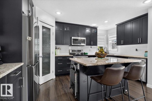 Modern kitchen featuring dark cabinetry, stainless steel appliances, a center island with bar seating, light-toned granite countertops, and wood-finish flooring - 13 Baie Madelene, Beaumont, AB 