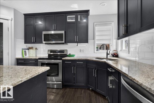 Modern kitchen featuring dark cabinetry, stainless steel appliances, speckled stone countertops, and subway tile backsplash - 13 Baie Madelene, Beaumont, AB 