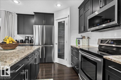 Kitchen featuring dark wood-finish cabinetry with silver hardware, granite-style countertops, stainless steel appliances, a white subway tile backsplash, and recessed lighting - 13 Baie Madelene, Beaumont, AB 