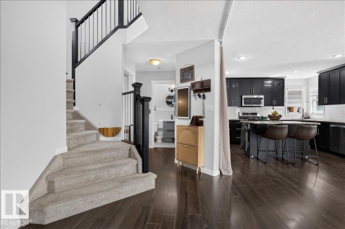 Staircase with carpeted treads and black balusters, dark wood-finish flooring, and a kitchen featuring black cabinetry, light countertops, and a white tile backsplash - 13 Baie Madelene, Beaumont, AB 