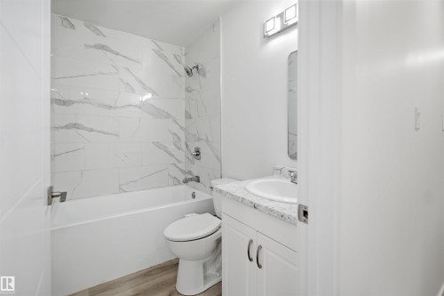 Modern bathroom featuring a white bathtub with a tile surround, a single-sink vanity with a light-colored countertop, and wood-finish flooring - 3548 20 Ave, Edmonton, AB 