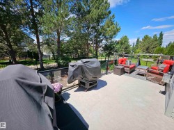 Spacious outdoor patio featuring concrete flooring, black metal railing with glass panels, and a backdrop of mature trees - 