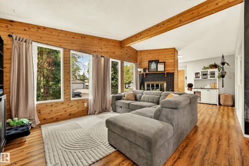 Spacious living area featuring wood-finish flooring, natural wood paneling, a row of large windows, exposed wood beams, and a black brick fireplace - 24 Addison Crescent, St. Albert, AB 