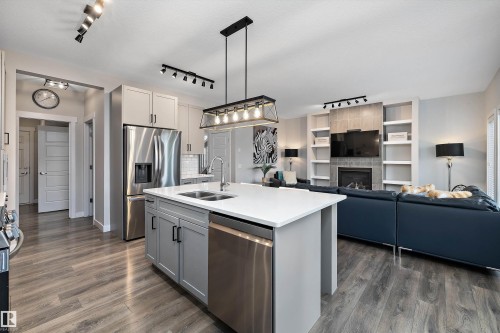 Open-concept living area featuring wood-finish flooring, a kitchen island with dual sink, stainless steel appliances, and a living room with a built-in fireplace and shelving - 363 Cy Becker Boulevard, Edmonton, AB - Indoor Photo Showing Kitchen With Fireplace With Double Sink