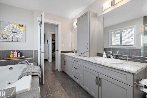 Bathroom featuring a large soaker tub with tiled surround, dual vanity with undermount sinks, stone-look countertops, and extensive gray cabinetry - 363 Cy Becker Boulevard, Edmonton, AB - Indoor Photo Showing Bathroom