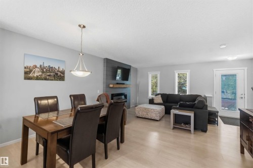 Open-concept living space featuring light wood-finish flooring, a contemporary fireplace with a dark paneled surround and floating mantel, and multiple windows providing natural light - 940 East Gate, Edmonton, AB 