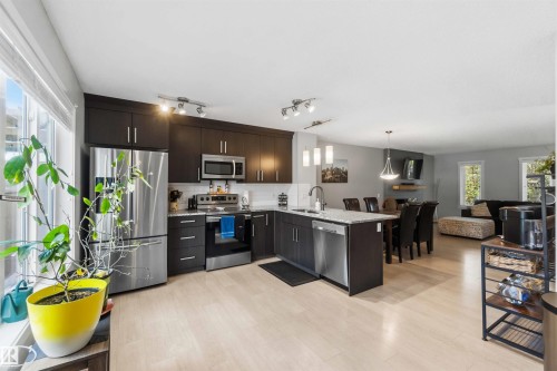 Open-concept kitchen featuring dark wood-finish cabinetry, stainless steel appliances, a center island with sink, and light wood-finish flooring - 940 East Gate, Edmonton, AB 