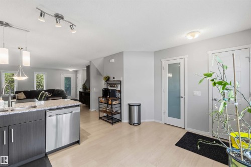 Open-concept living space with light wood-finish flooring, a kitchen island featuring dark cabinetry and light granite-style countertops, stainless steel appliances, and a modern track lighting fixture - 940 East Gate, Edmonton, AB 